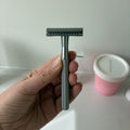 A hand holding a silver safety razor with a dark gray blade, set against a white bathroom background with a pink container visible.
