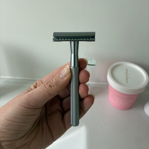 A hand holding a silver safety razor with a dark gray blade, set against a white bathroom background with a pink container visible.